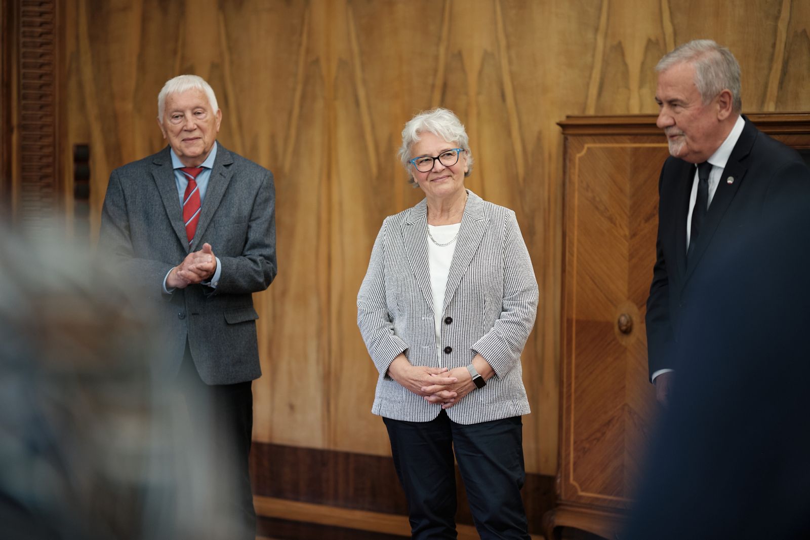 Trzy osoby stoją w reprezentacyjnym wnętrzu podczas oficjalnego spotkania, jedna z nich klaszcze; w tle drewniana ściana i meble.