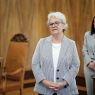 Osoba stoi w reprezentacyjnej sali i zwraca się do zgromadzonych; w tle widoczna jest inna osoba oraz drewniane ściany i meble.