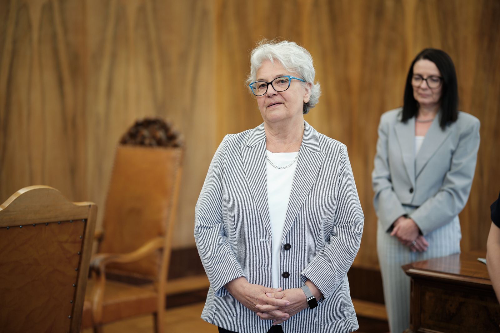 Osoba stoi w reprezentacyjnej sali i zwraca się do zgromadzonych; w tle widoczna jest inna osoba oraz drewniane ściany i meble.