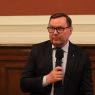 Osoba w garniturze siedzi przy stole i wypowiada się do mikrofonu podczas oficjalnego spotkania w sali z drewnianą boazerią.