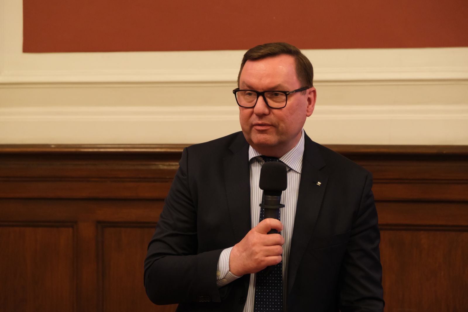 Osoba w garniturze siedzi przy stole i wypowiada się do mikrofonu podczas oficjalnego spotkania w sali z drewnianą boazerią.
