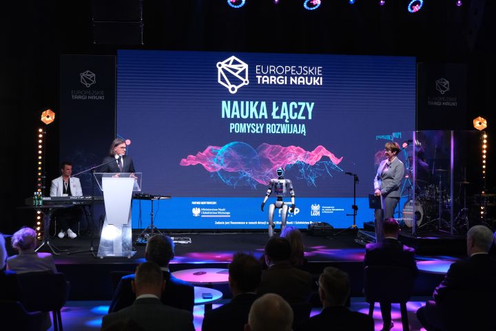 Osoba przemawia przy mównicy na scenie podczas wydarzenia „Europejskie Targi Nauki”, obok stoi humanoidalny robot; w tle widoczny jest duży ekran z hasłem „Nauka łączy, pomysły rozwijają” oraz publiczność.