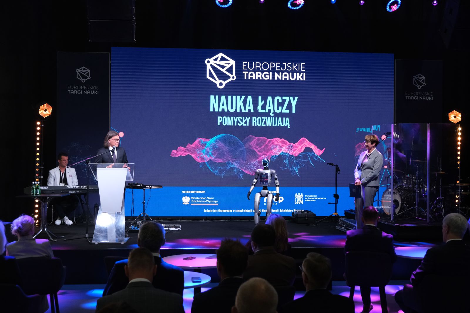 Osoba przemawia przy mównicy na scenie podczas wydarzenia „Europejskie Targi Nauki”, obok stoi humanoidalny robot; w tle widoczny jest duży ekran z hasłem „Nauka łączy, pomysły rozwijają” oraz publiczność.