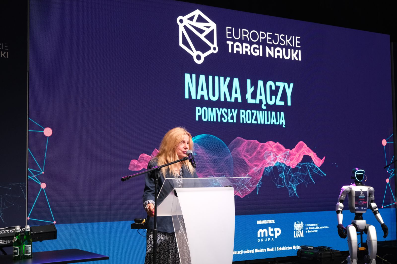 Osoba przemawia przy mównicy podczas wydarzenia „Europejskie Targi Nauki”; w tle widoczny jest duży ekran z napisem „Nauka łączy, pomysły rozwijają” oraz humanoidalny robot stojący obok sceny.