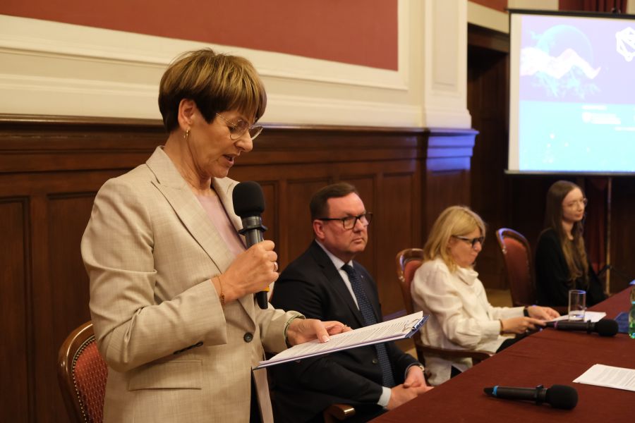 Osoba stoi przy stole i przemawia do zgromadzonych, trzymając mikrofon i kartkę z notatkami; w tle siedzą uczestnicy panelu oraz widoczny jest ekran prezentacji.