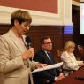 Osoba stoi przy stole i przemawia do zgromadzonych, trzymając mikrofon i kartkę z notatkami; w tle siedzą uczestnicy panelu oraz widoczny jest ekran prezentacji.