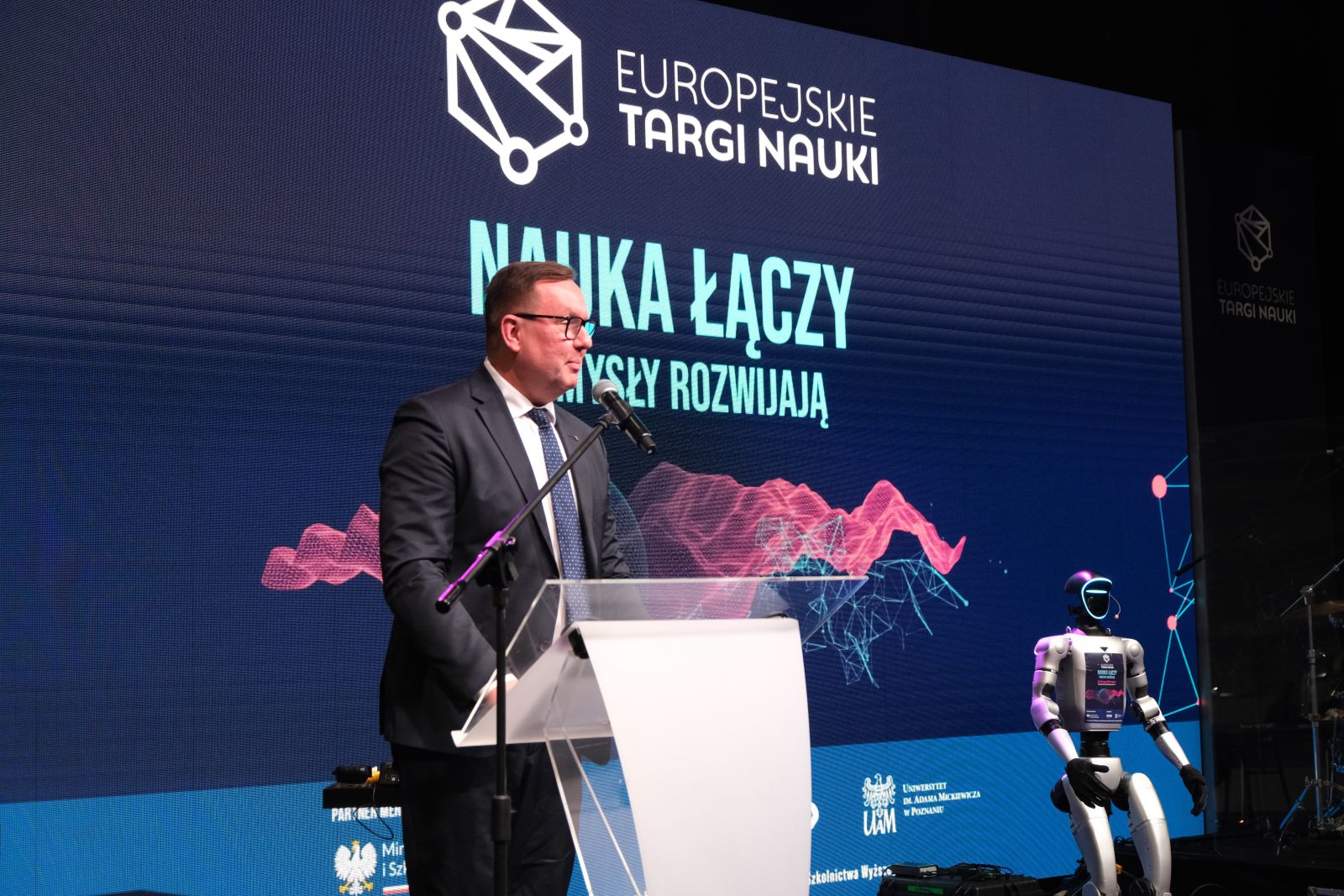 Osoba w garniturze przemawia do mikrofonu przy mównicy na tle ekranu z napisem „Europejskie Targi Nauki – Nauka łączy, pomysły rozwijają”; obok widoczny jest humanoidalny robot.