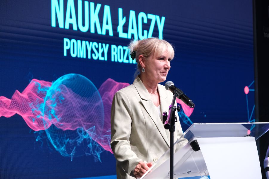 Zbliżenie na osobę przemawiającą do mikrofonu przy mównicy na tle ekranu z hasłem „Nauka łączy, pomysły rozwijają” i abstrakcyjną grafiką.