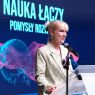 Zbliżenie na osobę przemawiającą do mikrofonu przy mównicy na tle ekranu z hasłem „Nauka łączy, pomysły rozwijają” i abstrakcyjną grafiką.