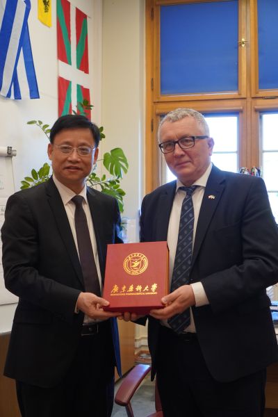 Dwie osoby stoją naprzeciw siebie i prezentują czerwone etui z napisem „Guangdong Pharmaceutical University”; w tle widoczne są flagi oraz okno w pomieszczeniu biurowym.