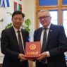 Dwie osoby stoją naprzeciw siebie i prezentują czerwone etui z napisem „Guangdong Pharmaceutical University”; w tle widoczne są flagi oraz okno w pomieszczeniu biurowym.