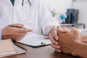 A doctor in a white gown with a stethoscope is interviewing a patient at a desk; there are notebooks and documents on the table.