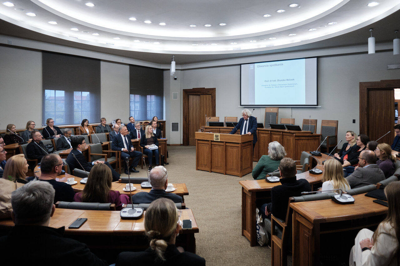 Uczestnicy uroczystości siedzący w sali Uniwersytetu im. Adama Mickiewicza w Poznaniu. Przy mównicy stoi mężczyzna przemawiający, w tle ekran z napisem „Otwarcie spotkania”.
