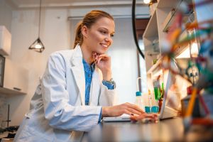 A smiling young researcher in a lab coat, staring at her laptop screen.