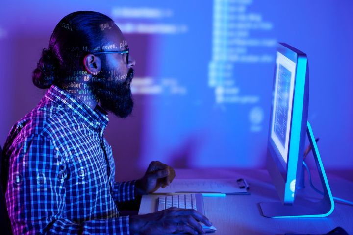 Mężczyzna pracujący przy komputerze, patrzący na ekran z widocznym kodem programistycznym, w pomieszczeniu o niebiesko-fioletowym oświetleniu.