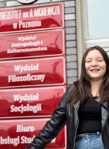 Uśmiechnięta młoda kobieta w czarnej skórzanej kurtce stoi przed czerwonymi tablicami informacyjnymi Uniwersytetu im. Adama Mickiewicza w Poznaniu, na których widnieją nazwy wydziałów, m.in. Wydziału Filozoficznego i Wydziału Socjologii.