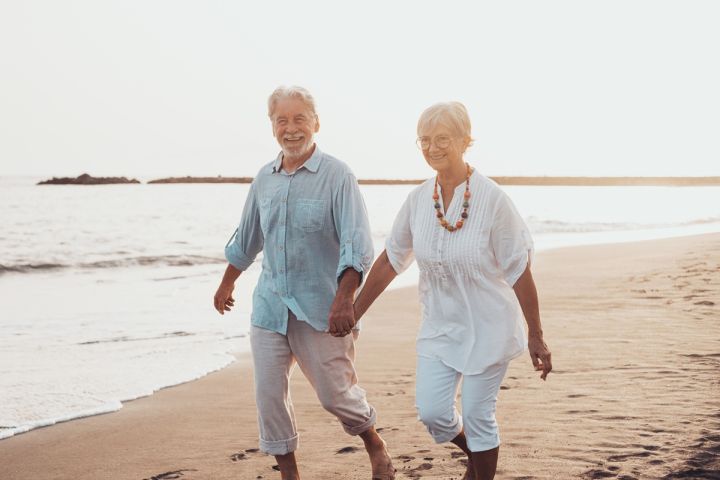 Uśmiechnięta para seniorów spaceruje boso po plaży, trzymając się za ręce.