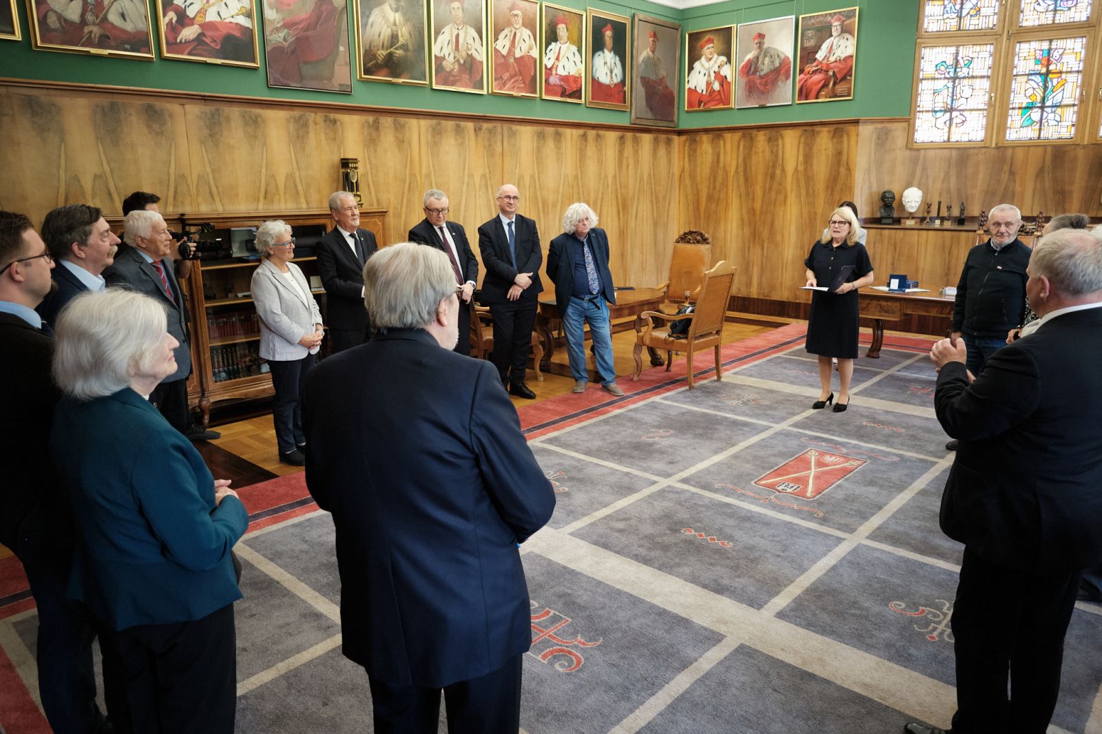 Grupa osób stoi w półokręgu w reprezentacyjnej sali z portretami na ścianach, słuchając osoby przemawiającej stojącej przy biurku.