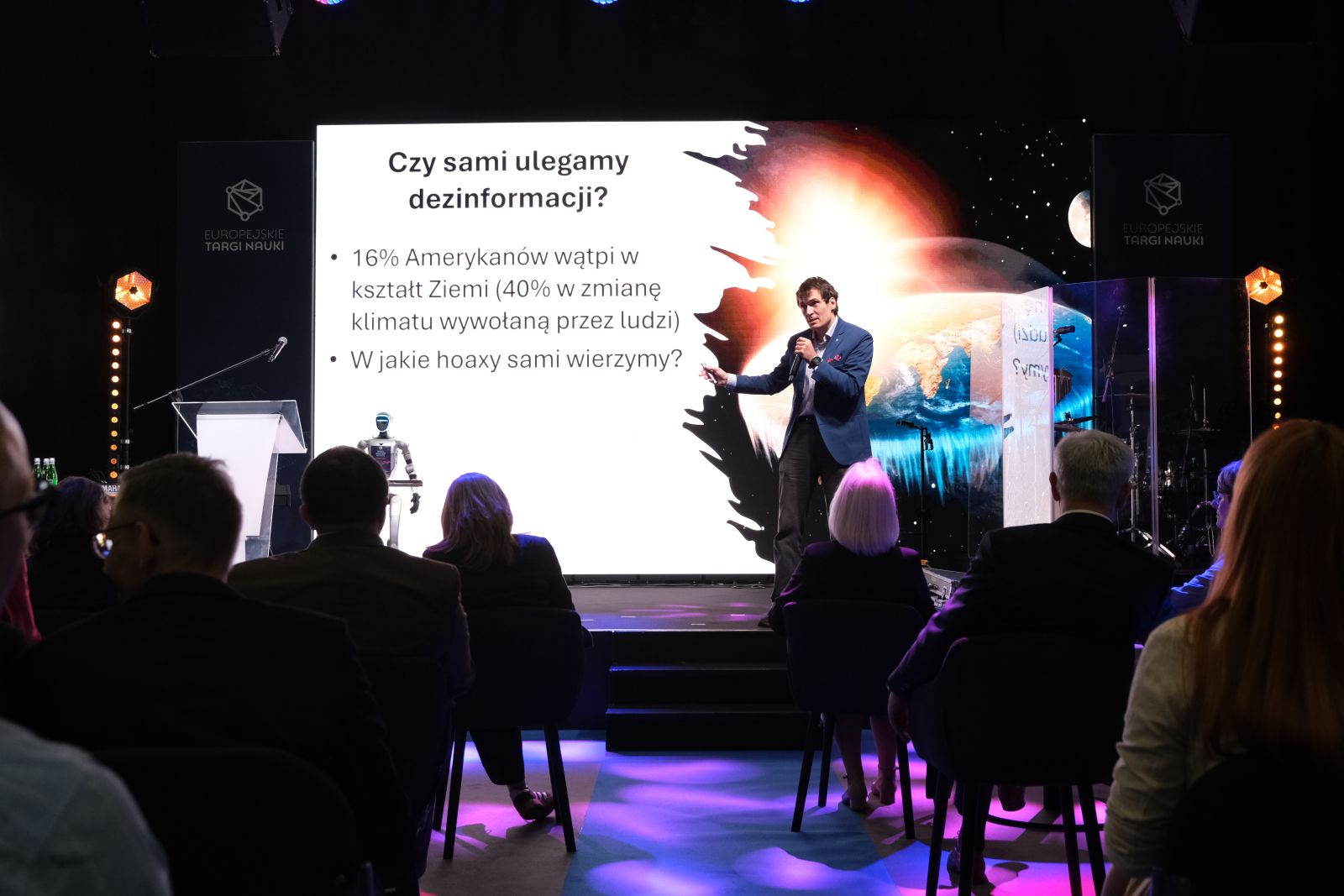 Prelegent stoi na scenie i prowadzi prezentację na temat dezinformacji; na dużym ekranie widoczny jest slajd z pytaniem „Czy sami ulegamy dezinformacji?” oraz grafika Ziemi, a przed sceną siedzi publiczność.