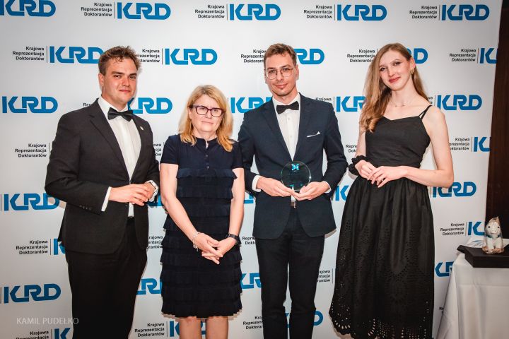 Cztery osoby stoją przed ścianką z logotypem „KRD – Krajowa Reprezentacja Doktorantów”; jedna osoba w smokingu trzyma statuetkę, obok stoją inni uczestnicy w formalnych strojach.