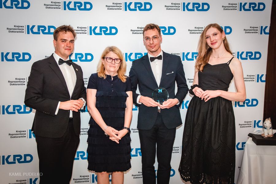 Cztery osoby stoją przed ścianką z logotypem „KRD – Krajowa Reprezentacja Doktorantów”; jedna osoba w smokingu trzyma statuetkę, obok stoją inni uczestnicy w formalnych strojach.