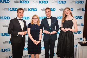 Cztery osoby stoją przed ścianką z logotypem „KRD – Krajowa Reprezentacja Doktorantów”; jedna osoba w smokingu trzyma statuetkę, obok stoją inni uczestnicy w formalnych strojach.