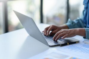 Zbliżenie na dłonie piszącej osoby pracującej na laptopie przy biurku; obok komputera leżą długopis i wydruki z wykresami.