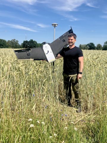 Mężczyzna stojący wśród wysokiego zboża trzyma duży dron o konstrukcji skrzydłowej. W tle błękitne niebo i drzewa.