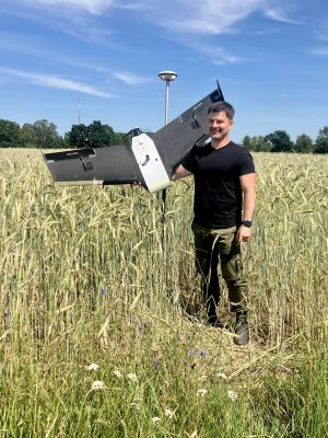 Mężczyzna stojący wśród wysokiego zboża trzyma duży dron o konstrukcji skrzydłowej. W tle błękitne niebo i drzewa.