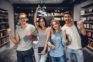 „Grupa czterech uśmiechniętych młodych osób z książkami i notatnikami, stojących w bibliotece i gestykulujących z entuzjazmem.