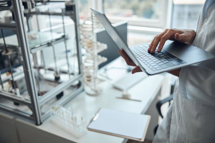  A person in a white lab coat is holding an open laptop in a laboratory, entering data; research equipment, test tubes and documents can be seen on the worktop.