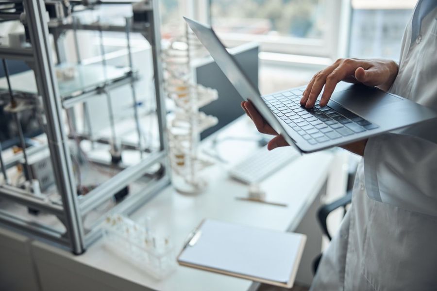  A person in a white lab coat is holding an open laptop in a laboratory, entering data; research equipment, test tubes and documents can be seen on the worktop.