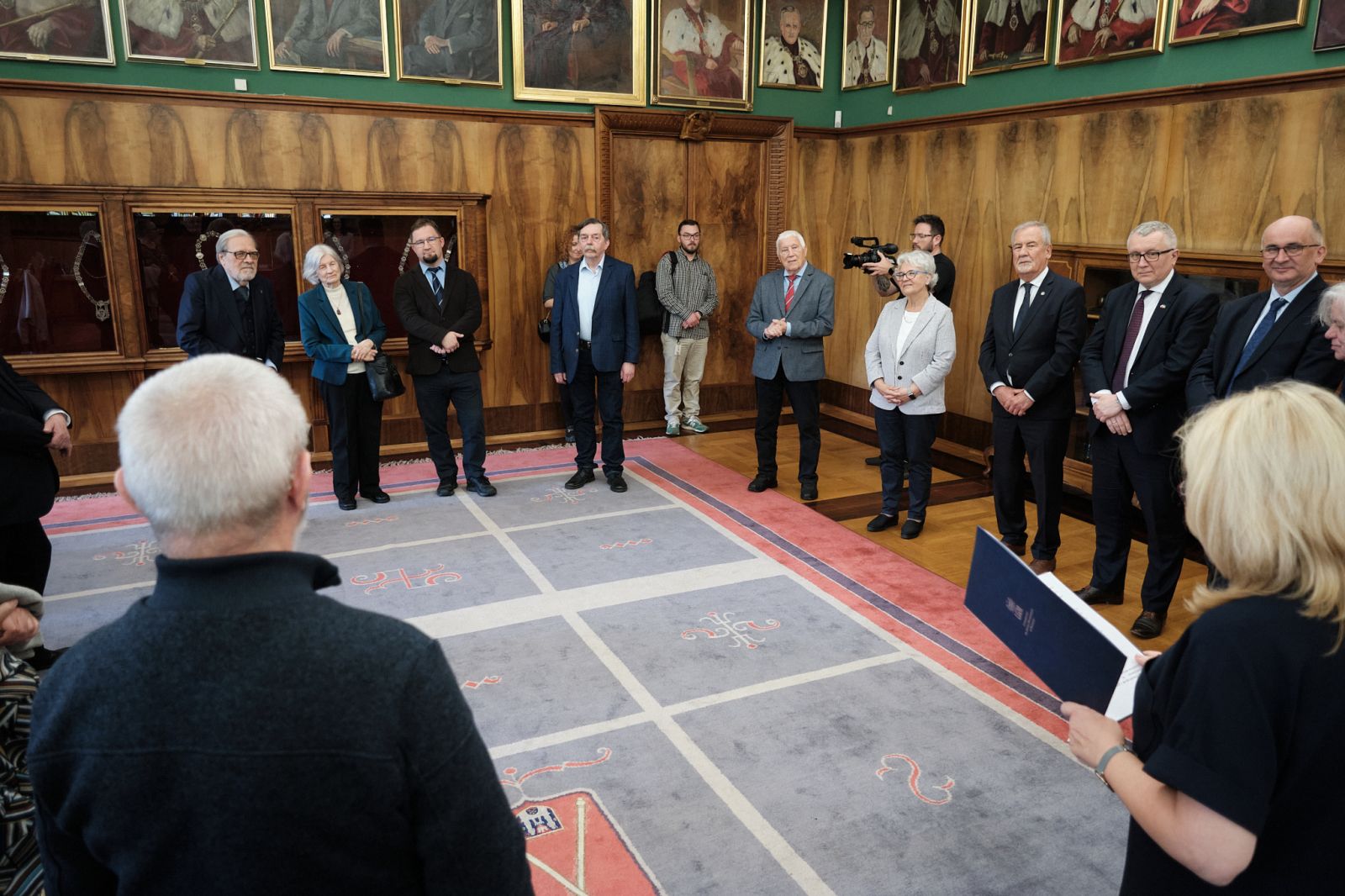 Uczestnicy wydarzenia stoją w eleganckim wnętrzu z drewnianymi ścianami i portretami, tworząc półokrąg wokół osoby trzymającej dokument.