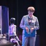 Osoba stoi na scenie obok humanoidalnego robota i trzyma teczkę z napisem UAM; w tle widoczne są elementy sceniczne oraz oświetlenie wydarzenia.