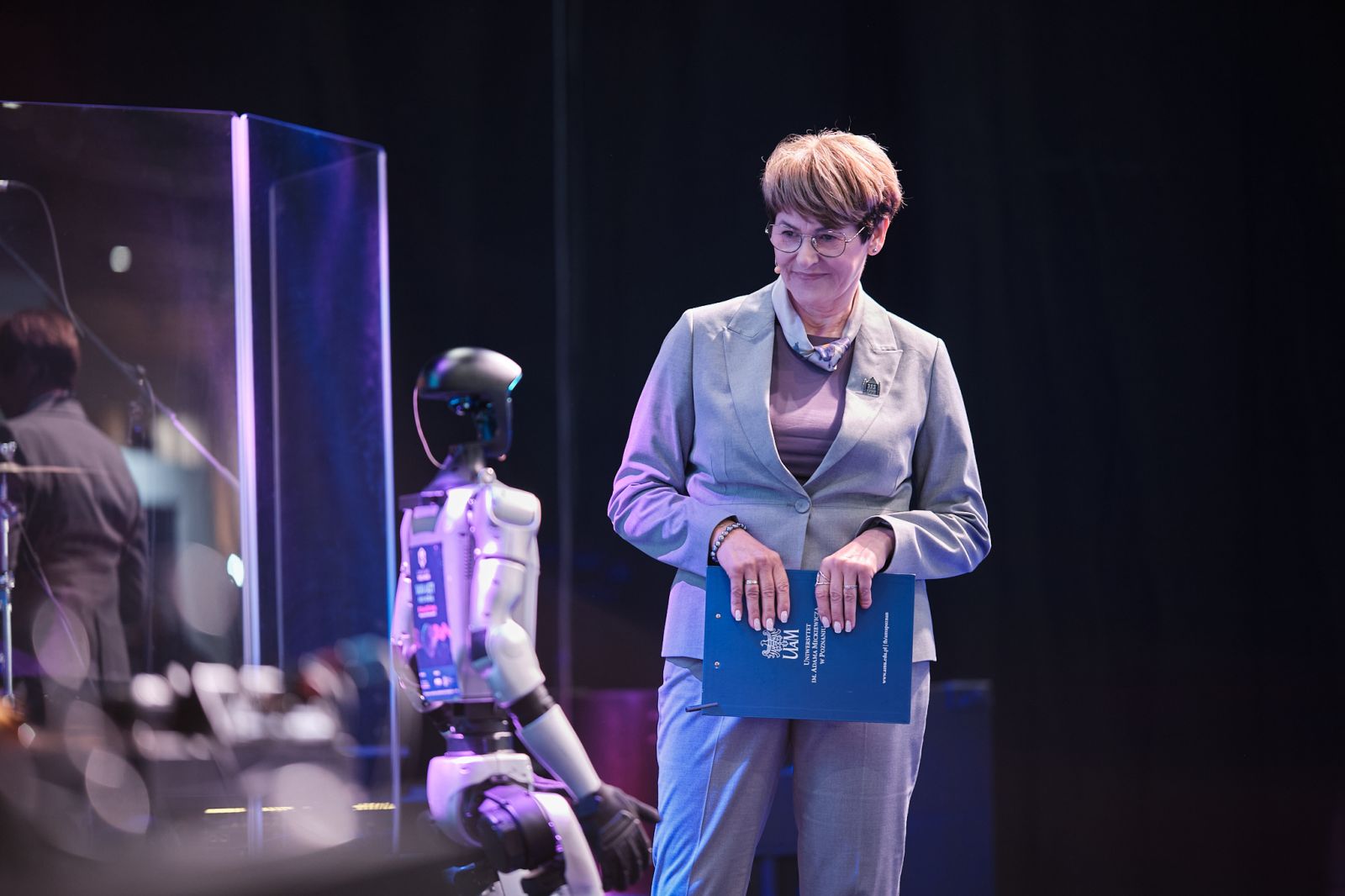 Osoba stoi na scenie obok humanoidalnego robota i trzyma teczkę z napisem UAM; w tle widoczne są elementy sceniczne oraz oświetlenie wydarzenia.