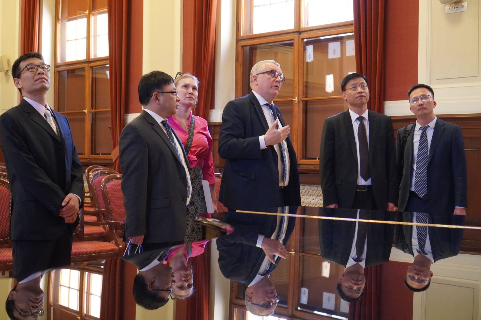 Grupa osób stoi wokół fortepianu w eleganckim wnętrzu z wysokimi oknami i czerwonymi zasłonami; jedna osoba gestykuluje podczas omawiania elementów sali.