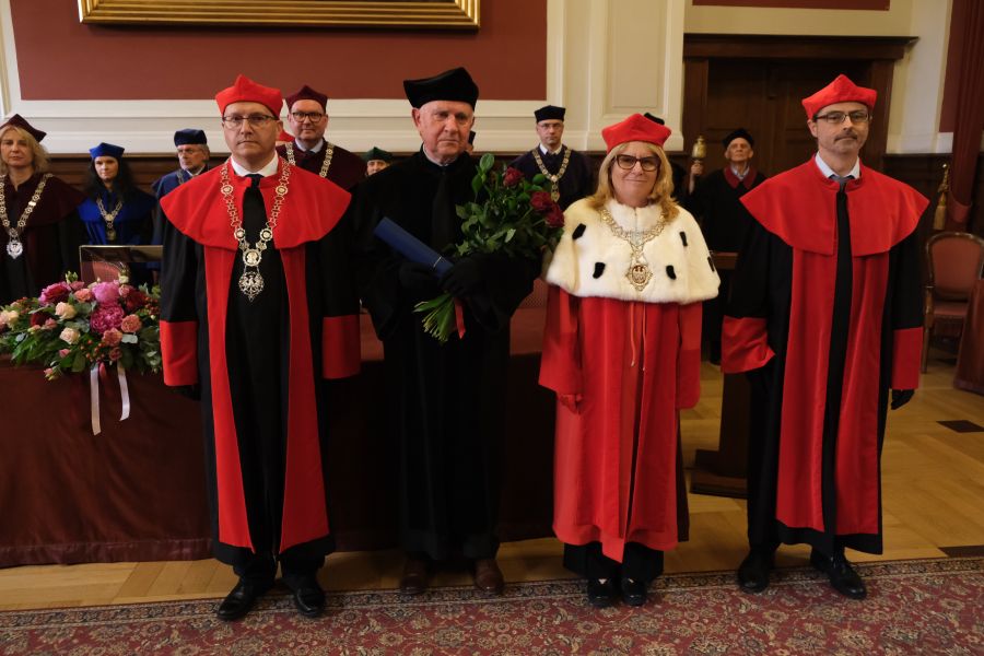 Grupa osób w togach akademickich stoi w rzędzie podczas uroczystości; w centrum osoba trzyma dyplom i bukiet róż, w tle widoczne są dekoracje kwiatowe i wnętrze sali.