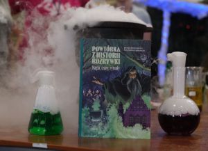 Książka „Powtórka z historii rozrywki. Magia, czary, rytuały” stoi na stole obok kolb laboratoryjnych z kolorowymi cieczami, z których unosi się para, tworząc efekt eksperymentu chemicznego.