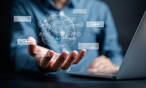 Dłoń trzymająca wirtualną kulę z napisem „AI” i ikonami technologii; w tle laptop.