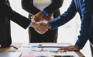 Uścisk dłoni dwóch osób w garniturach nad stołem z dokumentami podczas spotkania biznesowego.