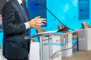 Mężczyzna w garniturze stoi przy przezroczystej mównicy z mikrofonem podczas wystąpienia lub konferencji.