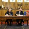 Dwie osoby siedzą przy dużym drewnianym biurku i podpisują dokumenty w reprezentacyjnym wnętrzu z herbem i oznaczeniem UAM w tle.