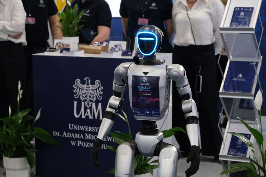 Zbliżenie na humanoidalnego robota stojącego przed stoiskiem Uniwersytetu im. Adama Mickiewicza; w tle widoczne są materiały promocyjne i osoby przy stoisku targowym.