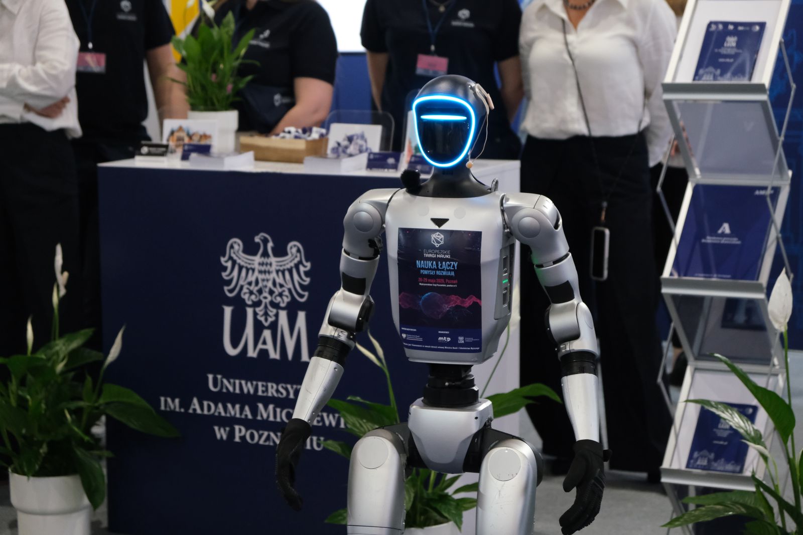 Zbliżenie na humanoidalnego robota stojącego przed stoiskiem Uniwersytetu im. Adama Mickiewicza; w tle widoczne są materiały promocyjne i osoby przy stoisku targowym.