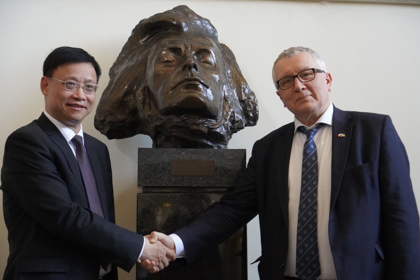 Dwie osoby w garniturach podają sobie ręce na tle popiersia rzeźby ustawionego na postumencie wewnątrz budynku.