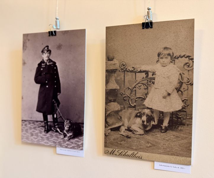 Dwie archiwalne fotografie prezentowane na wystawie przedstawiają dzieci w towarzystwie psów; zdjęcia zawieszone są na ścianie galerii wraz z podpisami identyfikującymi eksponaty.