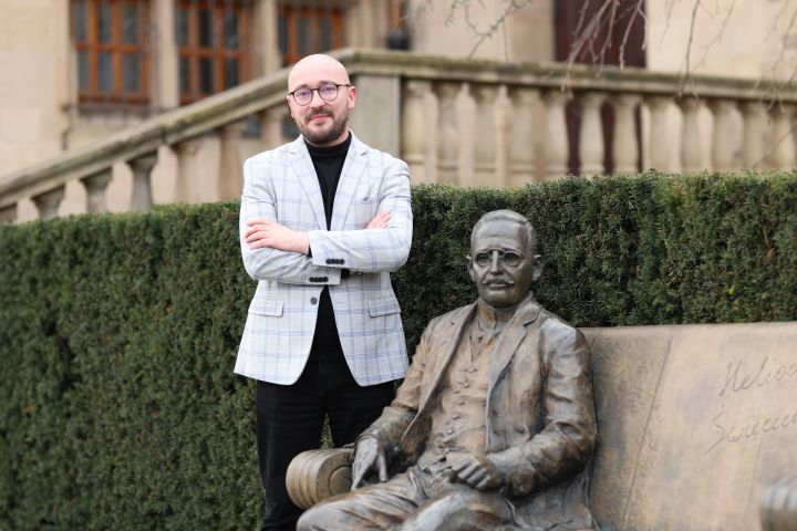 The graphic shows Dr Jakub Wojtczak standing by the Heliodor Święcicki bench, located in front of the Collegium Minus building. 