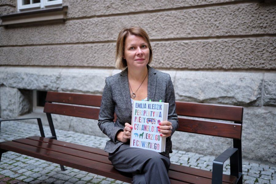 The pictures shows prof. Emilia Kledzik sitting on the bench in front of the AMU Faculty of Polish and Classical Philology. She holds her awarded book in her hands. 