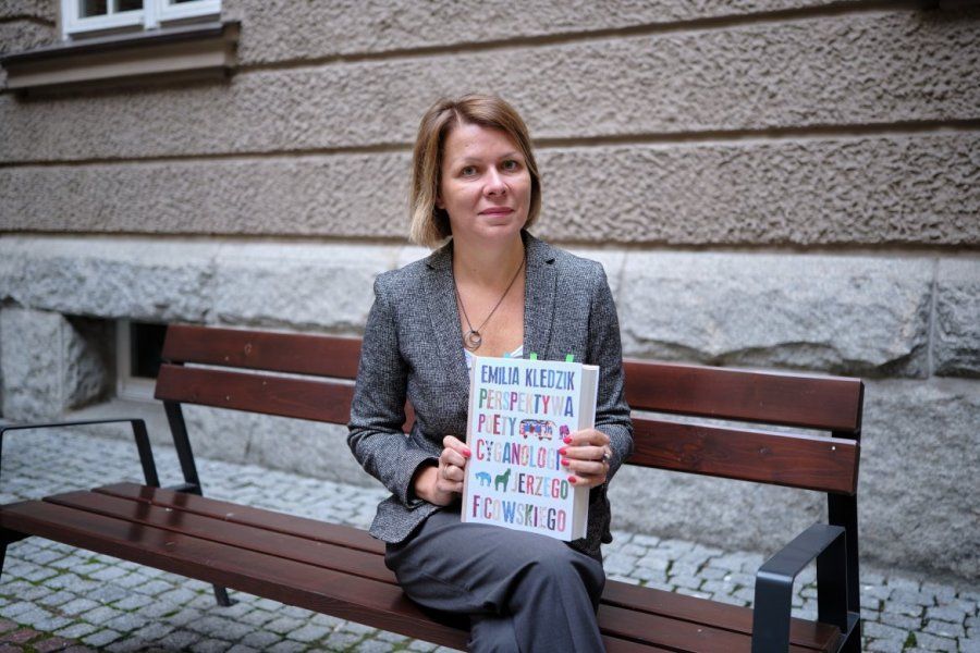 The pictures shows prof. Emilia Kledzik sitting on the bench in front of the AMU Faculty of Polish and Classical Philology. She holds her awarded book in her hands. 