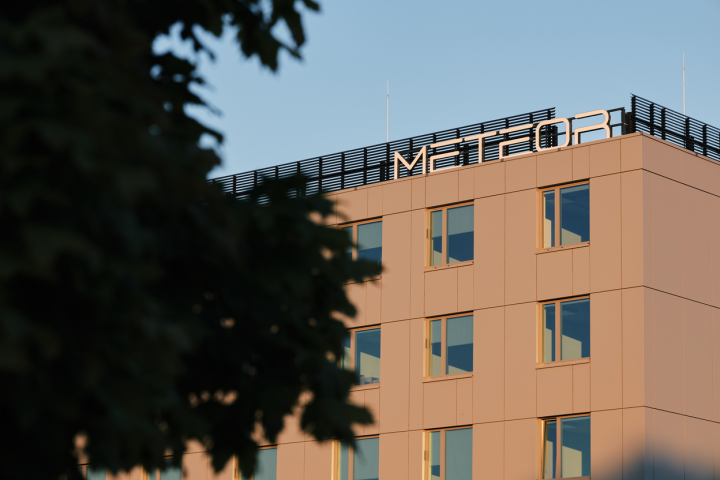 The picture shows the AMU Meteor Domitory at the time of a sunset. The building has a modern mass, painted in a light orange colour. At the top of the building the 